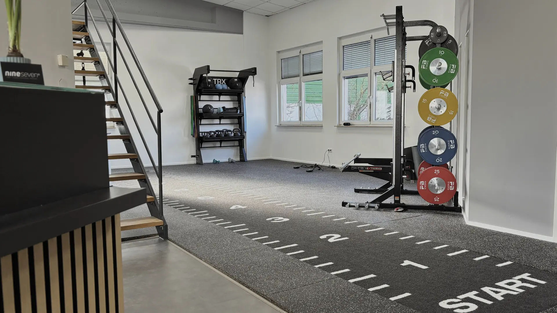 Das Bild zeigt das Fitnessstudio von nineseven Fitness in Ochsenfurt. Im Vordergrund ist eine Treppe zu sehen, die zu einem oberen Bereich führt. Rechts neben der Treppe befindet sich ein Regal mit TRX-Bändern, Medizinbällen und Kettlebells. An der rechten Wand ist eine Gewichtsscheibenhalterung mit farbigen Scheiben (10 kg grün, 15 kg gelb, 20 kg blau, 25 kg rot) angebracht. Der Boden ist mit einem Teppich ausgelegt, auf dem Markierungen und das Wort "START" zu sehen sind. Im Hintergrund sind zwei Fenster und weitere Trainingsgeräte zu erkennen. Das Bild ist im Querformat aufgenommen.