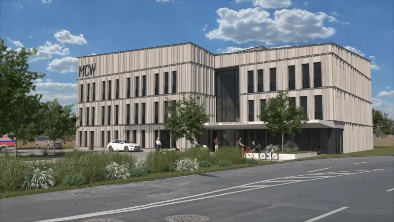 Das Bild zeigt das nineseven Fitnessstudio im Medical Center Windmühle in Unterpleichfeld von außen. Das Gebäude hat eine moderne Architektur mit einer grauen Fassade und großen Fenstern. Vor dem Eingang sind mehrere Personen zu sehen, die sich unterhalten oder das Gebäude betreten. Im Vordergrund sind gepflegte Grünflächen und ein Fahrradständer zu erkennen. Auf dem Gebäude ist das Logo „MCW“ zu sehen, und über dem Eingang steht „Center Unterpleichfeld“. Das Bild vermittelt einen Eindruck von der modernen und einladenden Atmosphäre des Fitnessstudios und des Medical Centers.