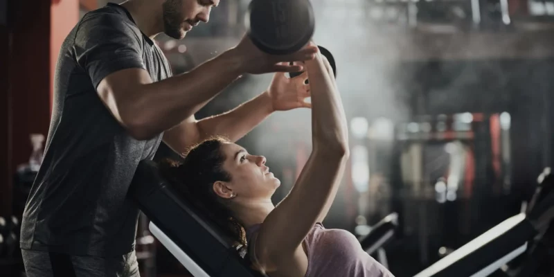 Das querformatige Bild zeigt eine Personal-Training-Situation in einem Fitnessstudio. Eine Person trägt ein lila Tanktop und liegt auf einer Schrägbank, während sie eine 15-kg-Hantel hebt. Eine andere Person, gekleidet in ein graues T-Shirt, steht daneben und bietet Unterstützung beim Training. Im Hintergrund sind verschiedene Trainingsgeräte erkennbar, die die professionelle Atmosphäre des Studios unterstreichen. Die Szene vermittelt den Eindruck von intensivem Training und persönlicher Betreuung durch einen Trainer.