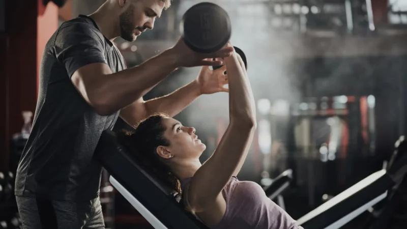 Das querformatige Bild zeigt eine Personal-Training-Situation in einem Fitnessstudio. Eine Person trägt ein lila Tanktop und liegt auf einer Schrägbank, während sie eine 15-kg-Hantel hebt. Eine andere Person, gekleidet in ein graues T-Shirt, steht daneben und bietet Unterstützung beim Training. Im Hintergrund sind verschiedene Trainingsgeräte erkennbar, die die professionelle Atmosphäre des Studios unterstreichen. Die Szene vermittelt den Eindruck von intensivem Training und persönlicher Betreuung durch einen Trainer.