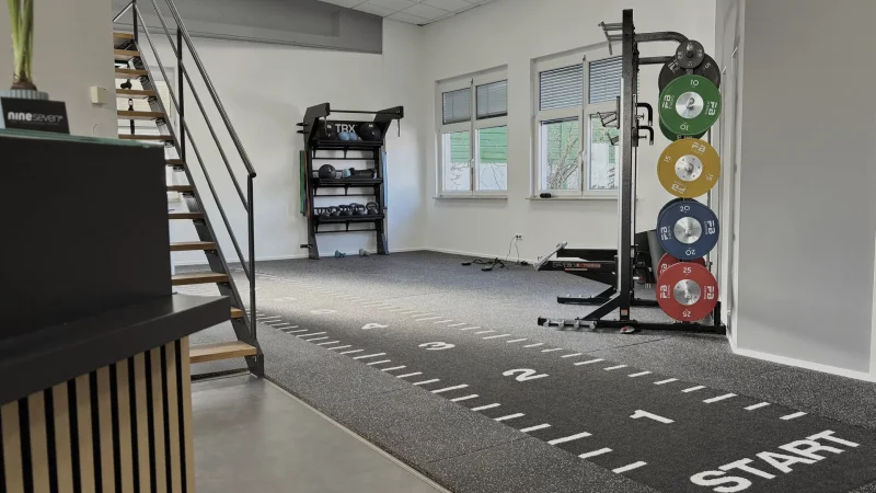 Das Bild zeigt das Fitnessstudio von nineseven Fitness in Ochsenfurt. Im Vordergrund ist eine Treppe zu sehen, die zu einem oberen Bereich führt. Rechts neben der Treppe befindet sich ein Regal mit TRX-Bändern, Medizinbällen und Kettlebells. An der rechten Wand ist eine Gewichtsscheibenhalterung mit farbigen Scheiben (10 kg grün, 15 kg gelb, 20 kg blau, 25 kg rot) angebracht. Der Boden ist mit einem Teppich ausgelegt, auf dem Markierungen und das Wort "START" zu sehen sind. Im Hintergrund sind zwei Fenster und weitere Trainingsgeräte zu erkennen. Das Bild ist im Querformat aufgenommen.