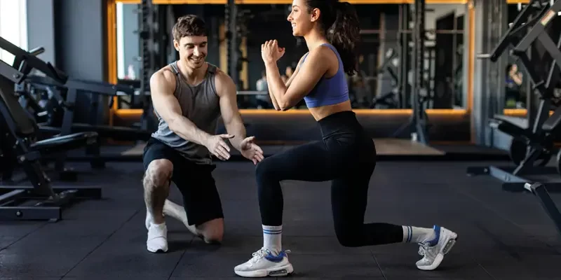 Das Bild zeigt zwei Personen in einem Fitnessstudio. Eine Person trägt ein blaues Sportoberteil und schwarze Leggings und führt eine Kniebeuge aus. Die andere Person trägt ein graues Tanktop und schwarze Shorts und kniet auf dem Boden, um Anweisungen zu geben. Im Hintergrund sind verschiedene Fitnessgeräte zu sehen. Das Bild stellt eine Trainingssituation dar und betont die Interaktion zwischen Trainer und Trainierendem.